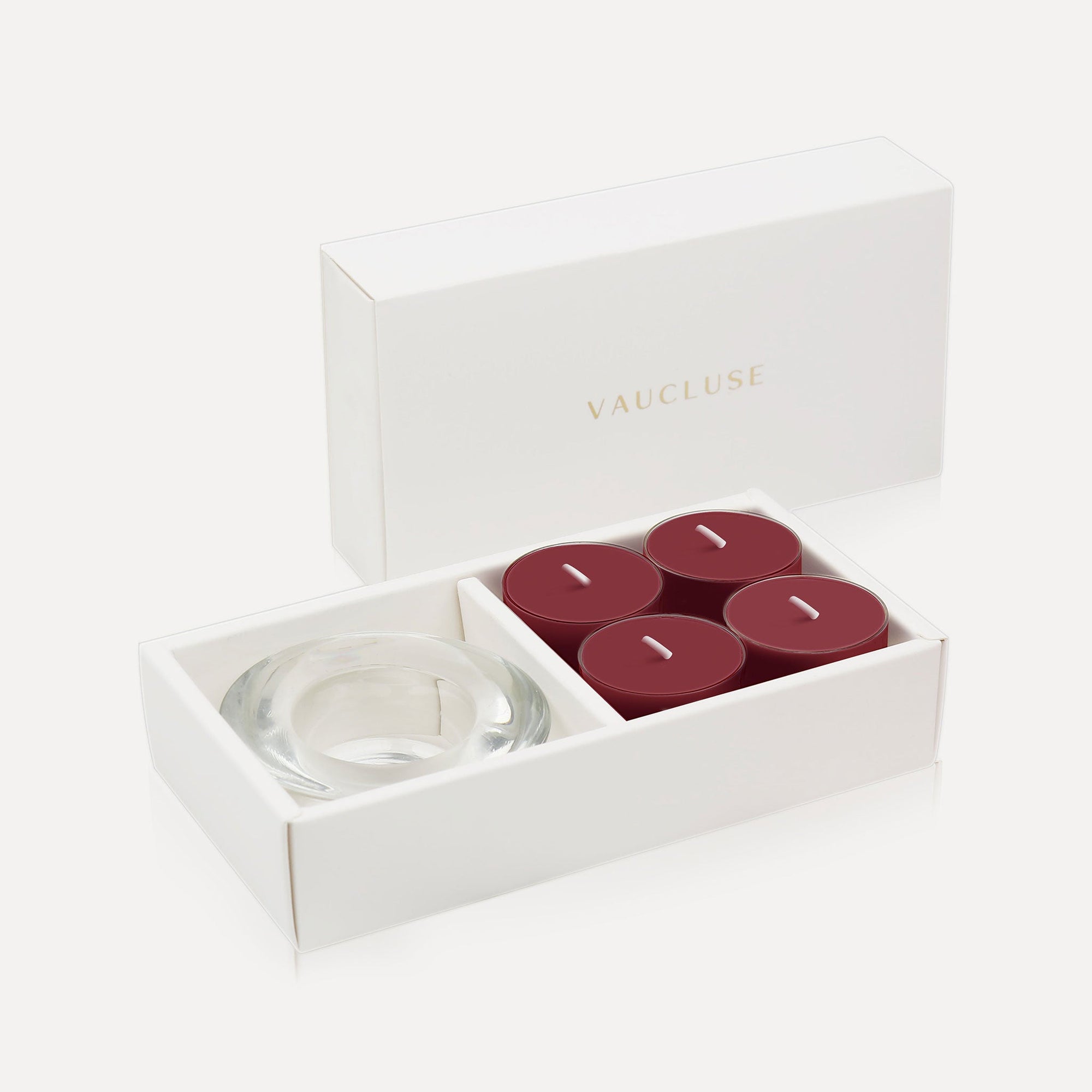 An open luxury white gift box displaying a set of four deep plum burgundy blackberry tealight candles next to a round clear glass candle holder.