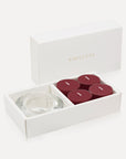 An open luxury white gift box displaying a set of four deep plum burgundy blackberry tealight candles next to a round clear glass candle holder.