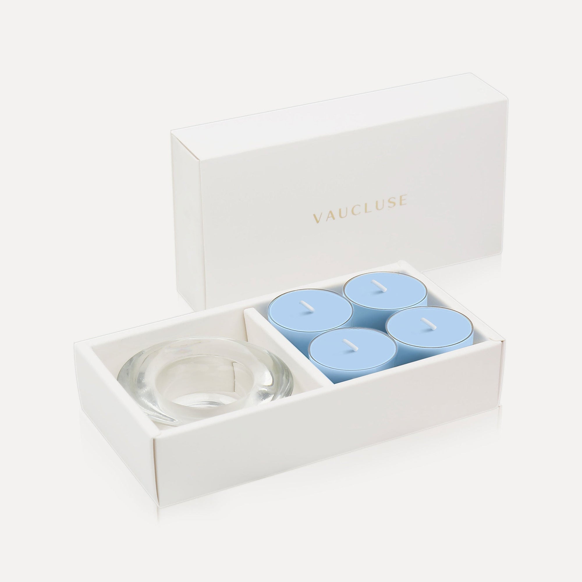 An open luxury white gift box displaying a set of four light blue breeze tealight candles next to a round clear glass candle holder.