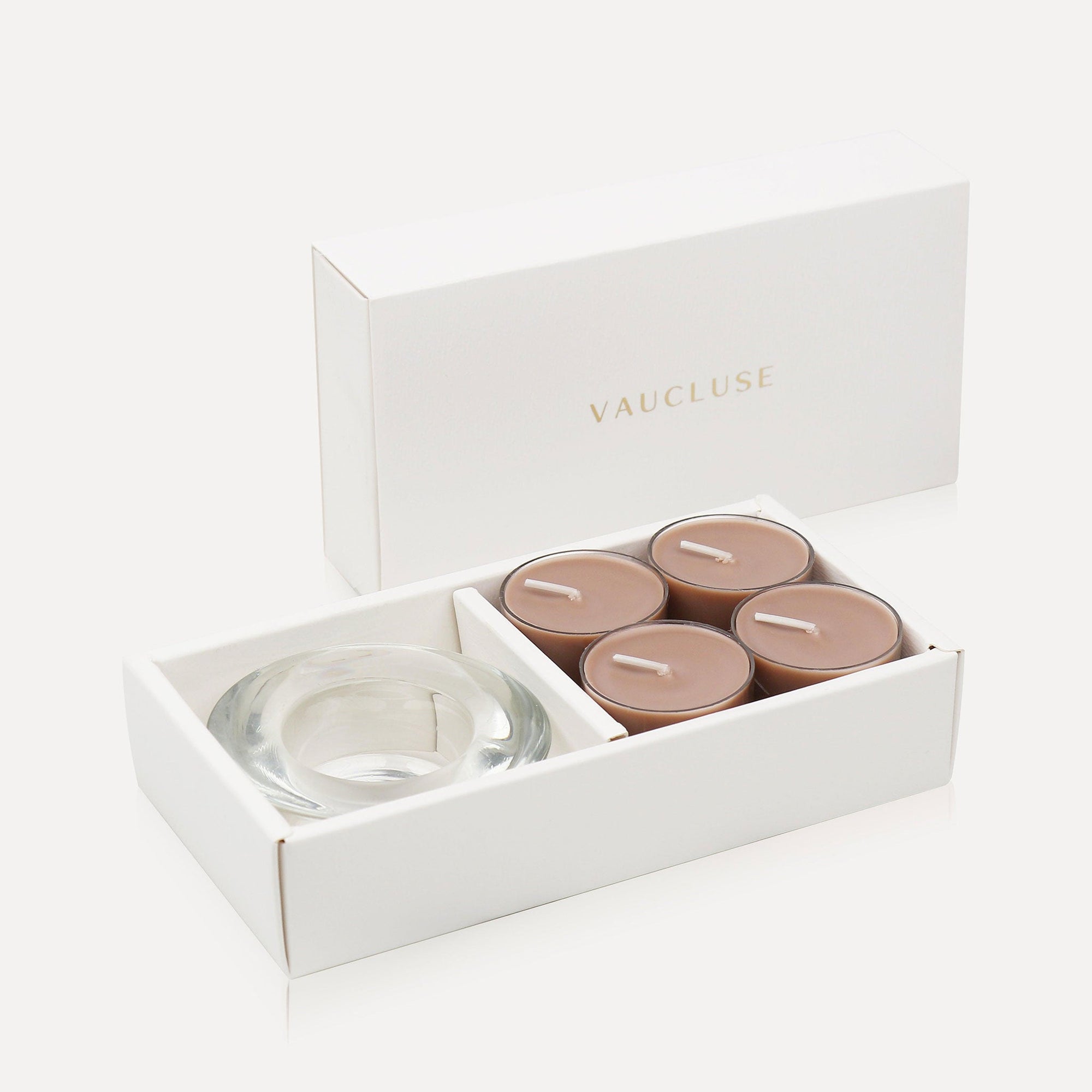 An open luxury white gift box cleanly displaying a set of four brown-hued cinnamon tealight candles alongside a thick round clear glass candle holder.