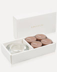 An open luxury white gift box cleanly displaying a set of four brown-hued cinnamon tealight candles alongside a thick round clear glass candle holder.