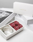 Premium retail unboxing presentation featuring the open burgundy blackberry tealight set resting against stacked white luxury gift boxes with gold branding.