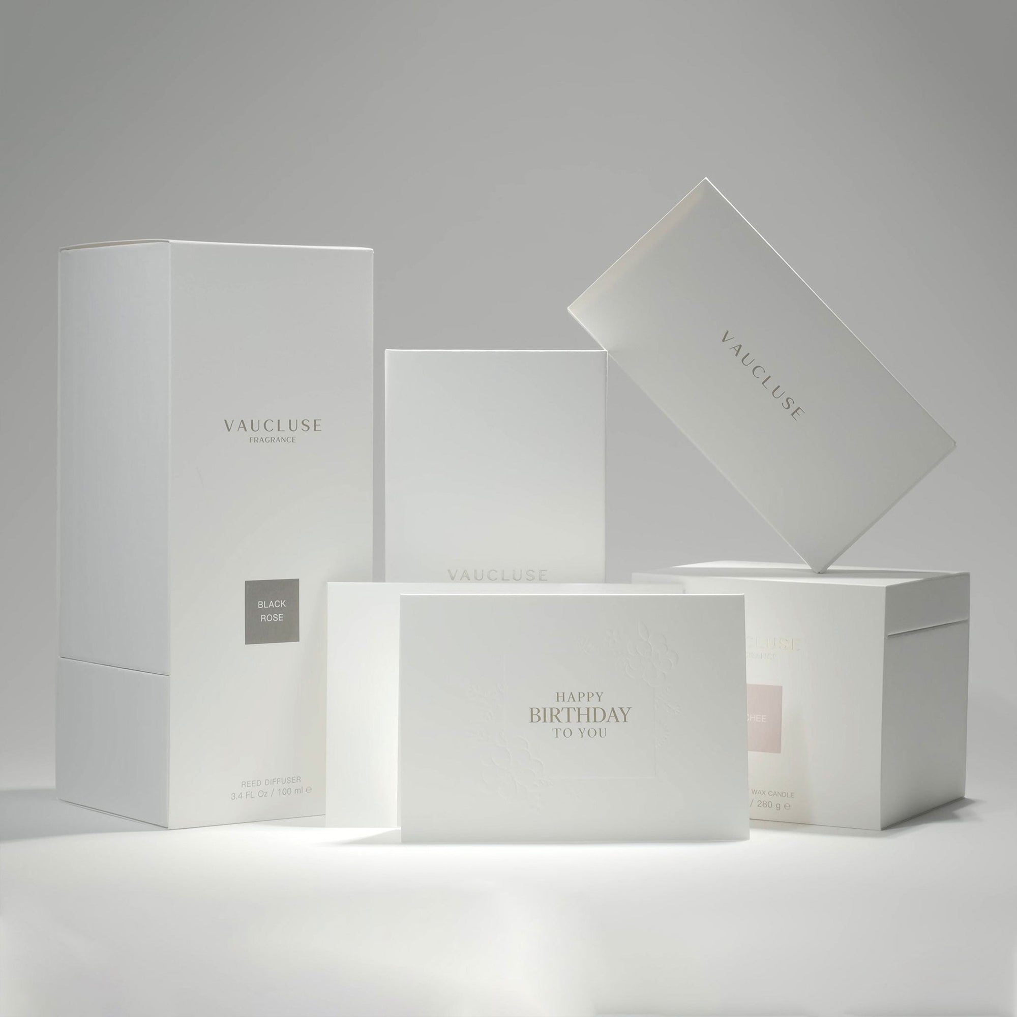 A curated composition of sleek white Vaucluse fragrance boxes, premium gift packaging, and the minimalist luxury birthday greeting card arranged together.