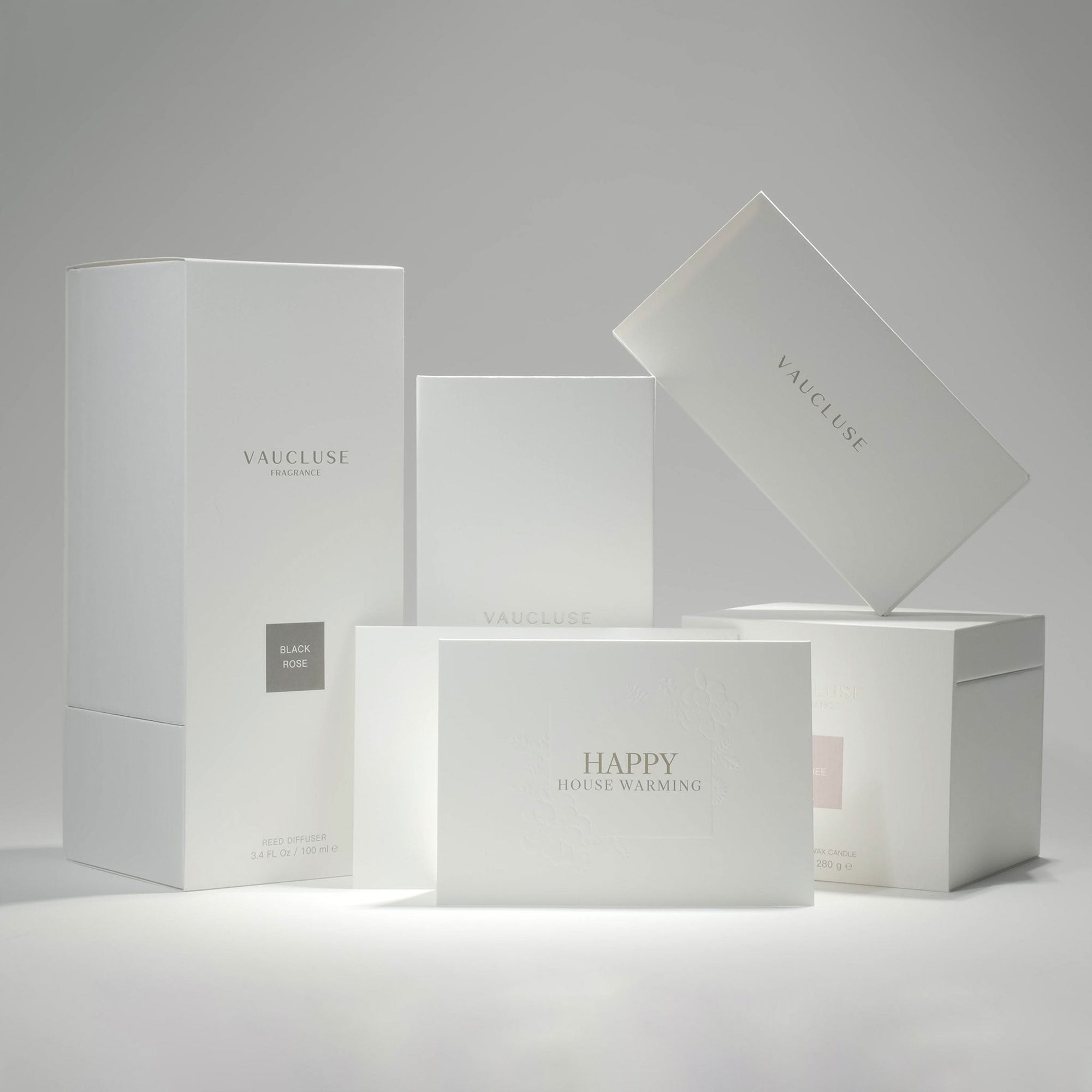 A curated collection of premium minimalist white Vaucluse fragrance boxes, candle packaging, and the elegant Happy House Warming card arranged neatly on a crisp white surface.