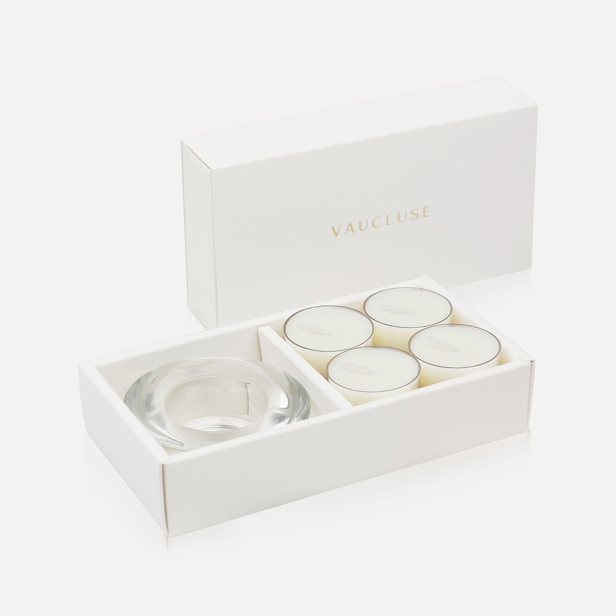 An open luxury white gift box displaying a set of four creamy white jasmine tealight candles next to a round clear glass candle holder.