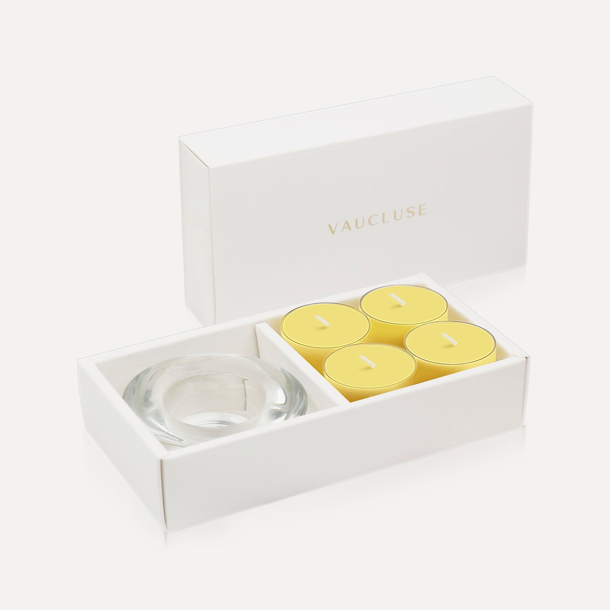 An open luxury white gift box displaying a set of four bright yellow lemon tealight candles next to a round clear glass candle holder.