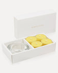 An open luxury white gift box displaying a set of four bright yellow lemon tealight candles next to a round clear glass candle holder.