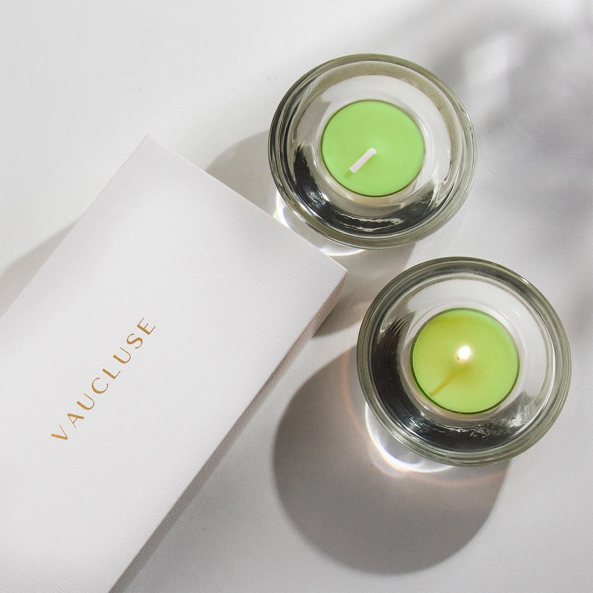 An overhead top-down view of two light green lily tealight candles in modern clear glass holders resting next to a white embossed Vaucluse gift box.