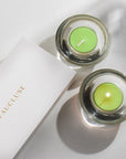 An overhead top-down view of two light green lily tealight candles in modern clear glass holders resting next to a white embossed Vaucluse gift box.