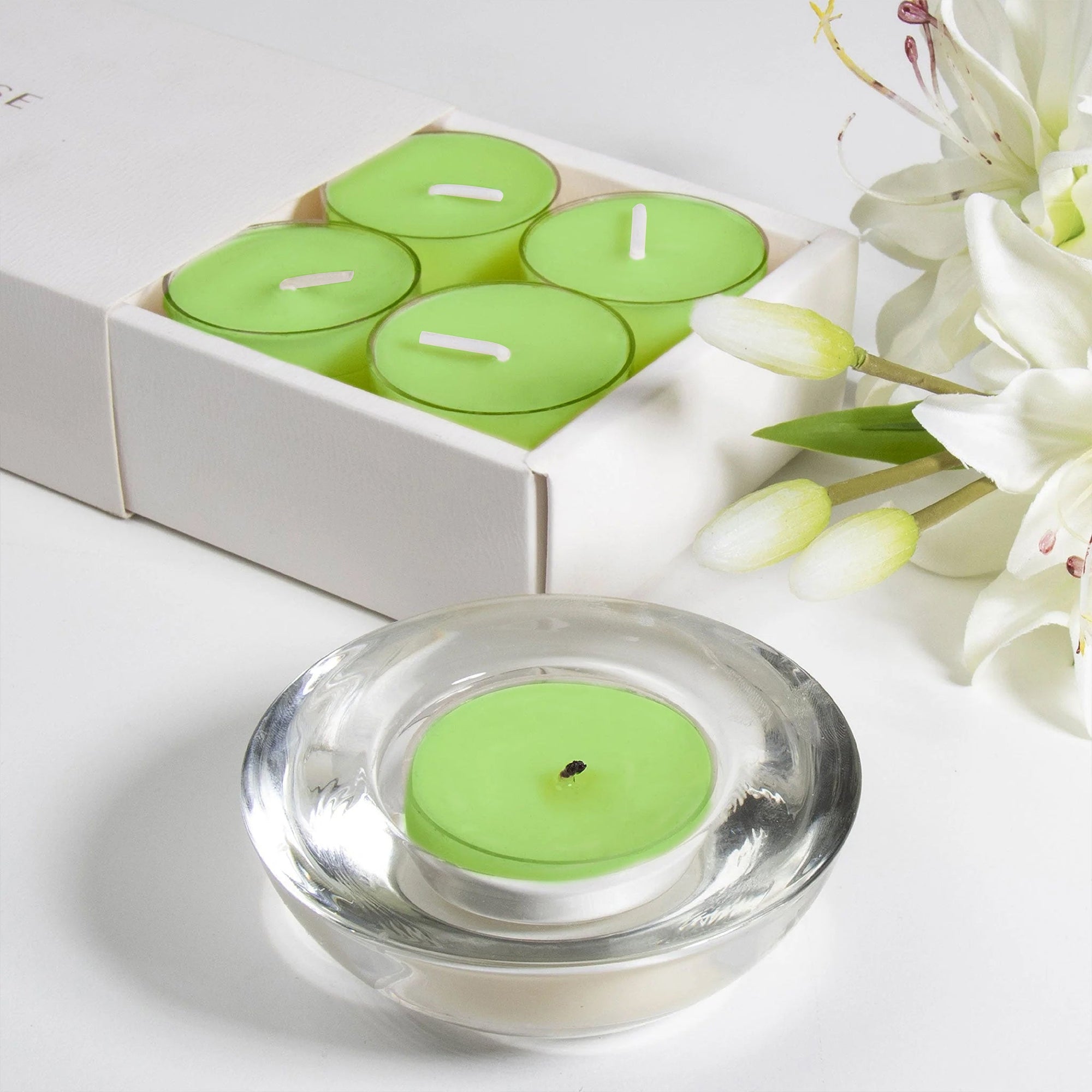 A light green lily scented tealight candle in a clear glass holder shown in the foreground with a partial view of the sliding box tray and white lilies.