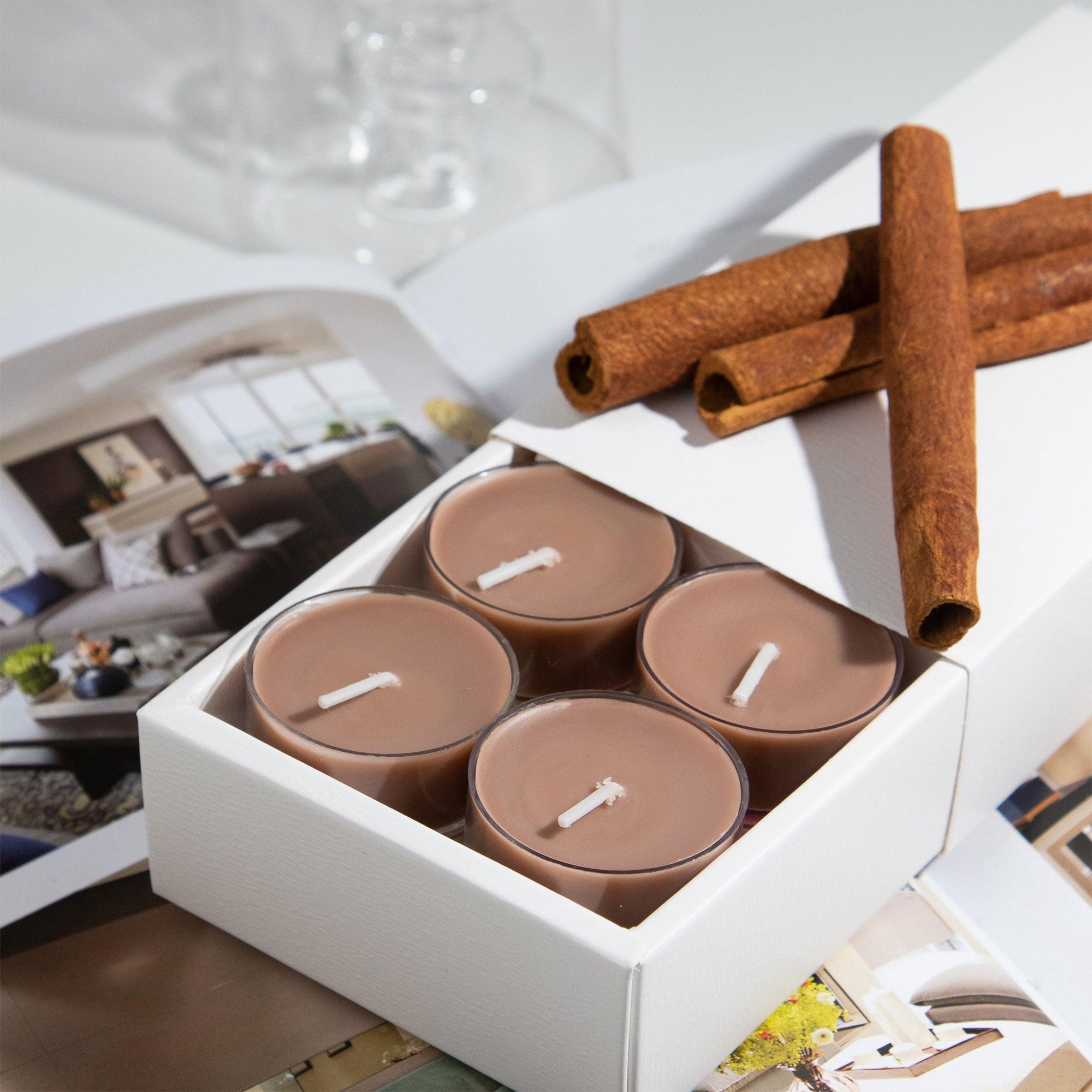 A small white box revealing four brown Vaucluse Cinnamon spiced tealights, arranged on an open magazine with decorative cinnamon sticks.