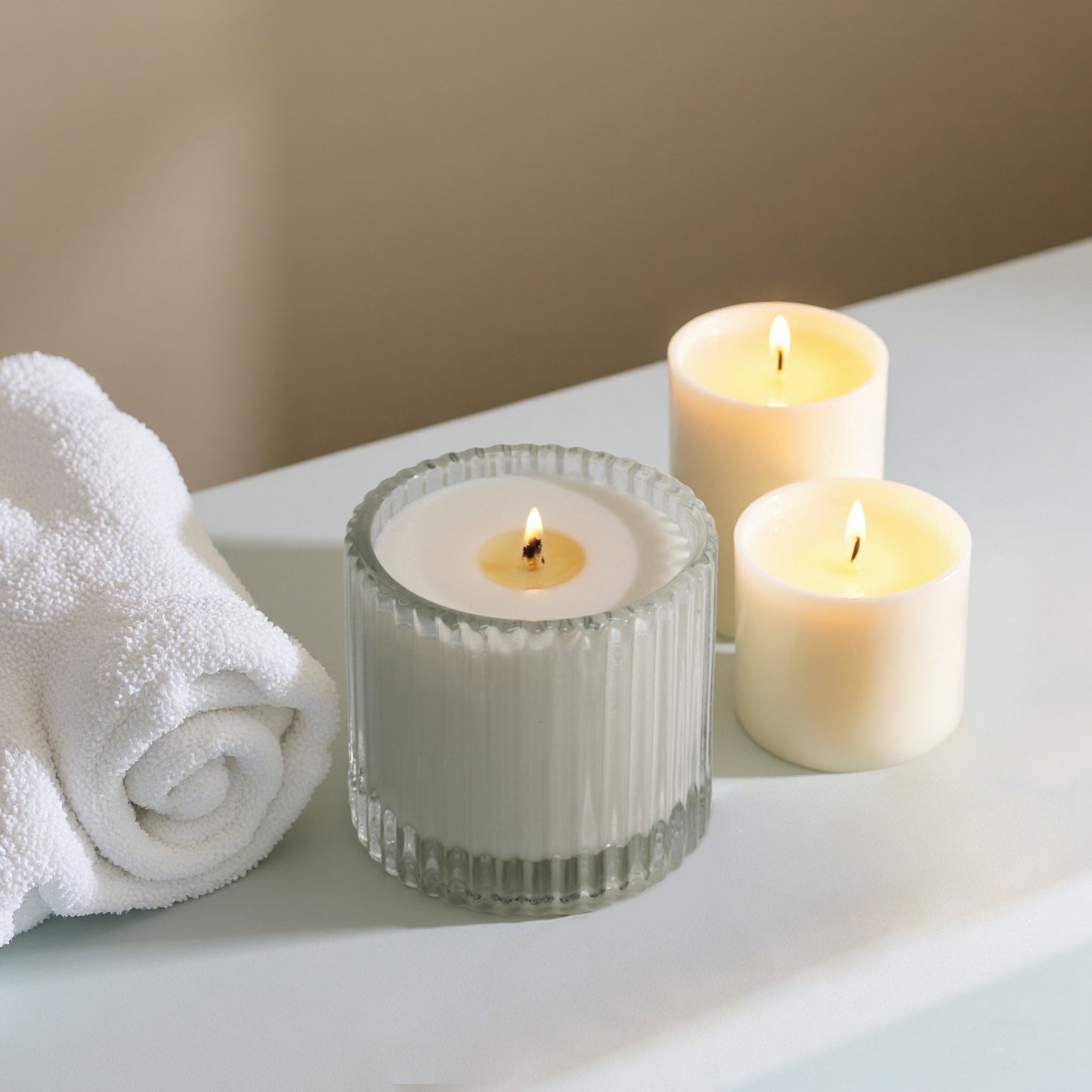 A ribbed glass Vaucluse candle alongside two smaller lit votive candles and a white rolled towel on a clean spa ledge.