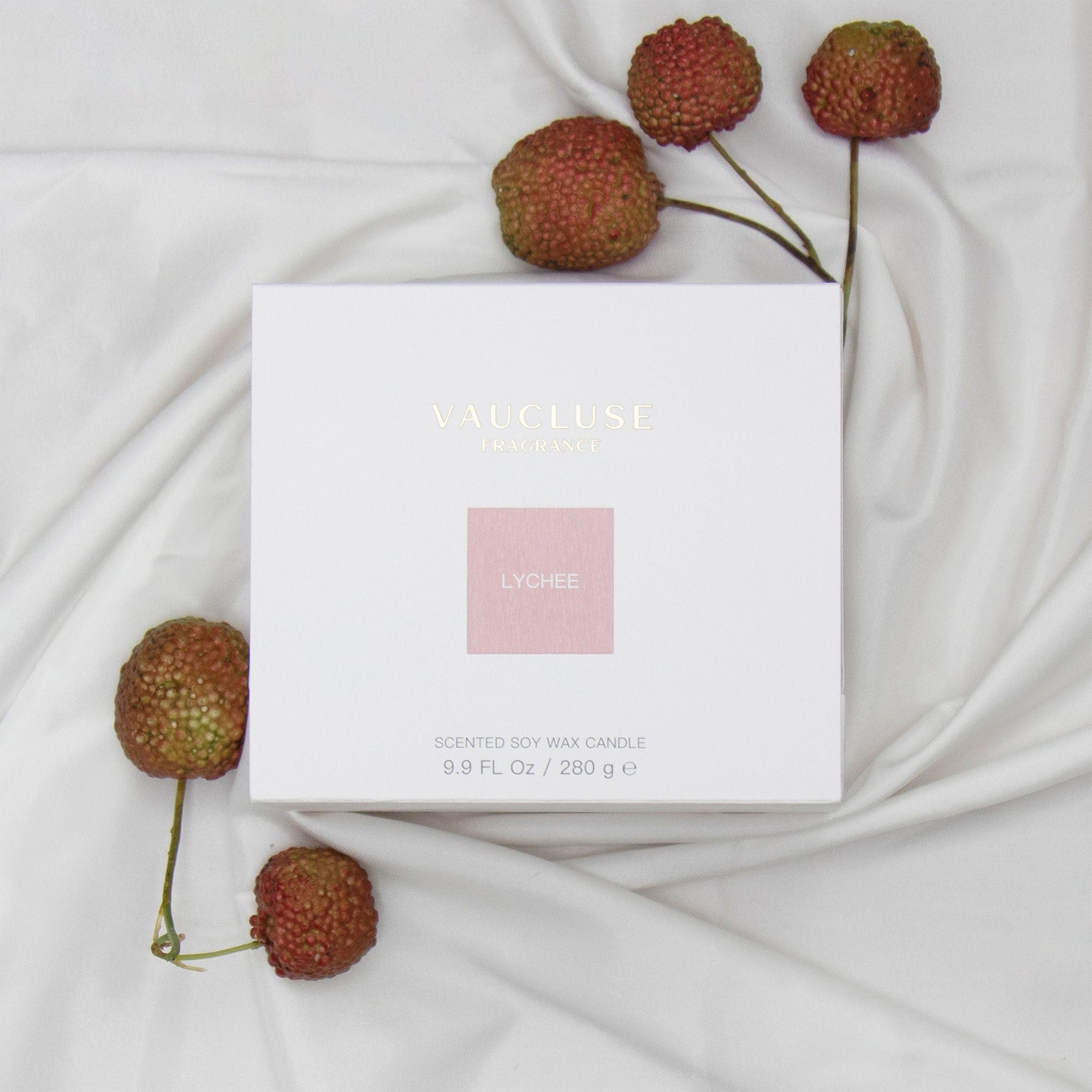 Top down view of the white square Vaucluse Lychee candle packaging box with a pink label, sitting on soft draped fabric next to fresh lychees on stems.