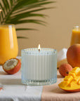 A lit Vaucluse Lychee luxury scented candle glass jar surrounded by fresh sliced mangoes, lychees, and tropical juice glasses.