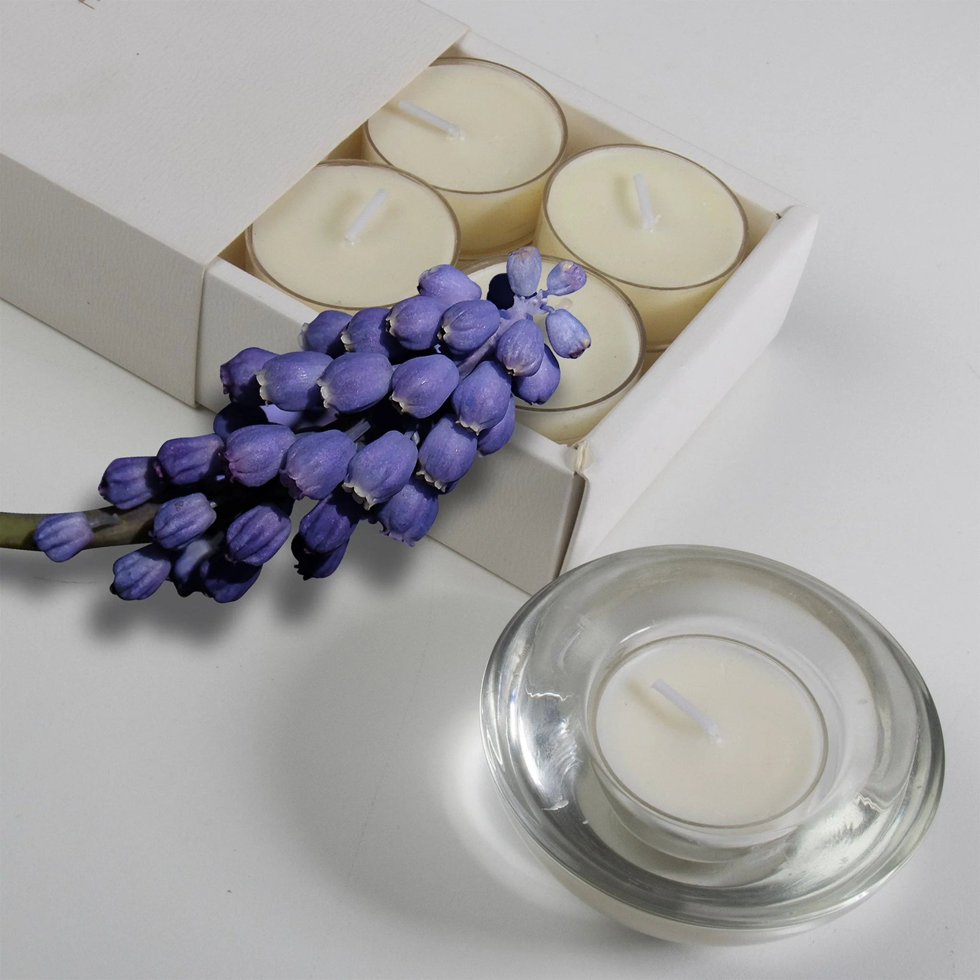 A cream musk scented tealight candle in a clear glass holder shown in the foreground with purple flowers resting on a partial view of the sliding box tray.