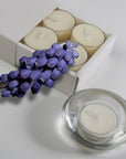 A cream musk scented tealight candle in a clear glass holder shown in the foreground with purple flowers resting on a partial view of the sliding box tray.