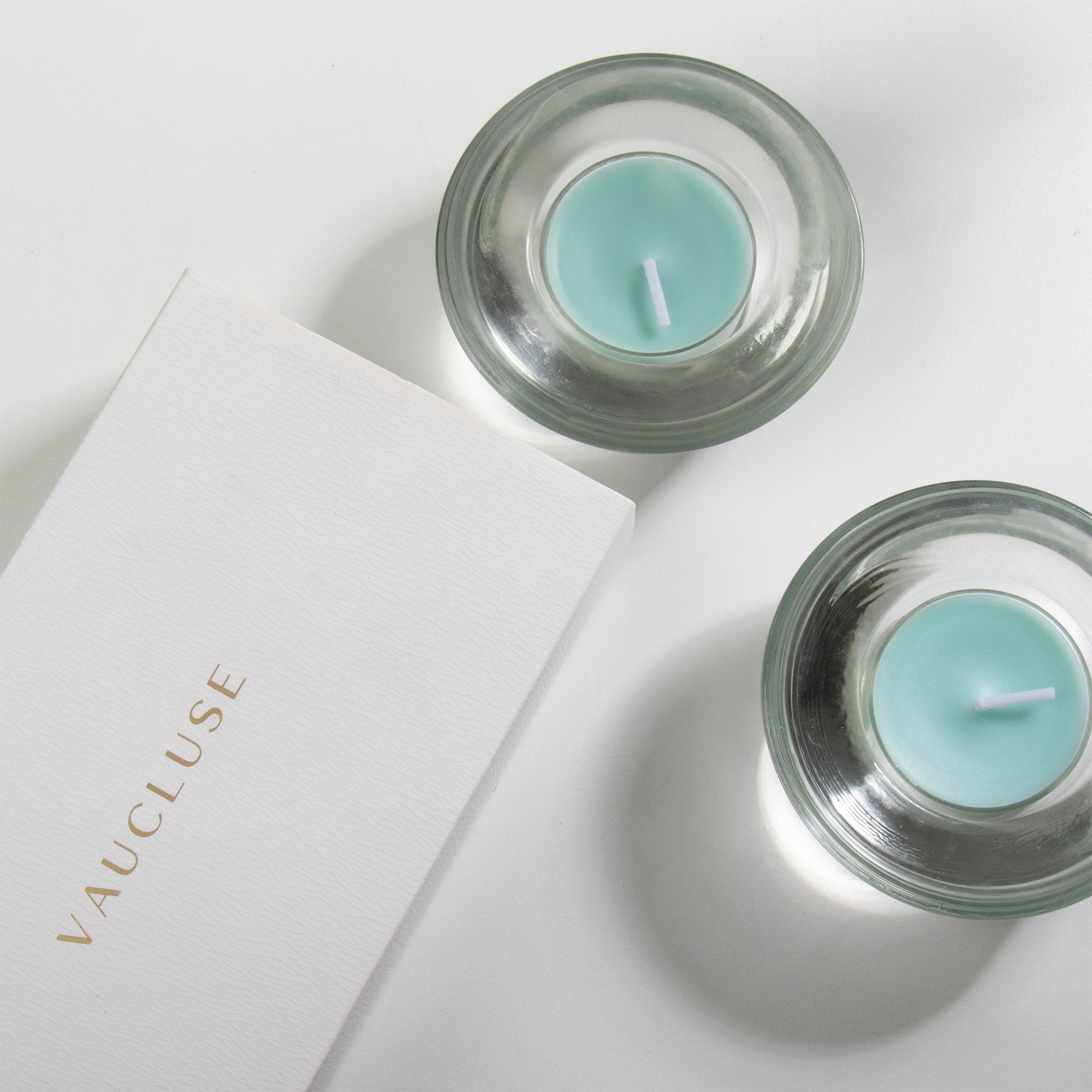 An overhead top-down view of teal ocean scented tealight candles resting in elegant clear glass holders next to a gold-embossed white Vaucluse gift box.