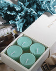 Four unlit light teal ocean scented tealight candles nestled inside a white branded Vaucluse gift box styled with deep blue coral texture in the background.