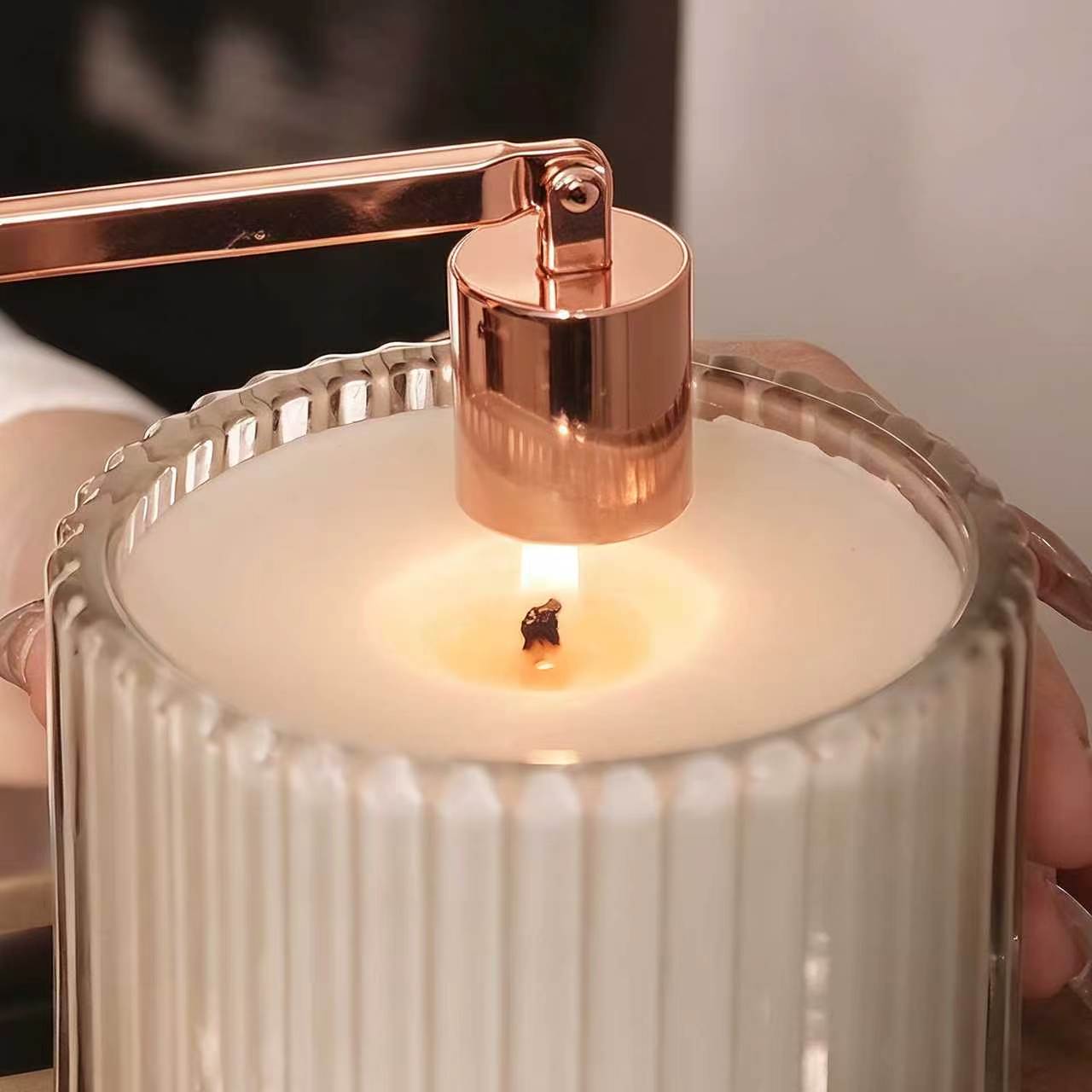 Using a rose gold candle snuffer to safely extinguish a burning luxury candle in a ribbed glass jar.