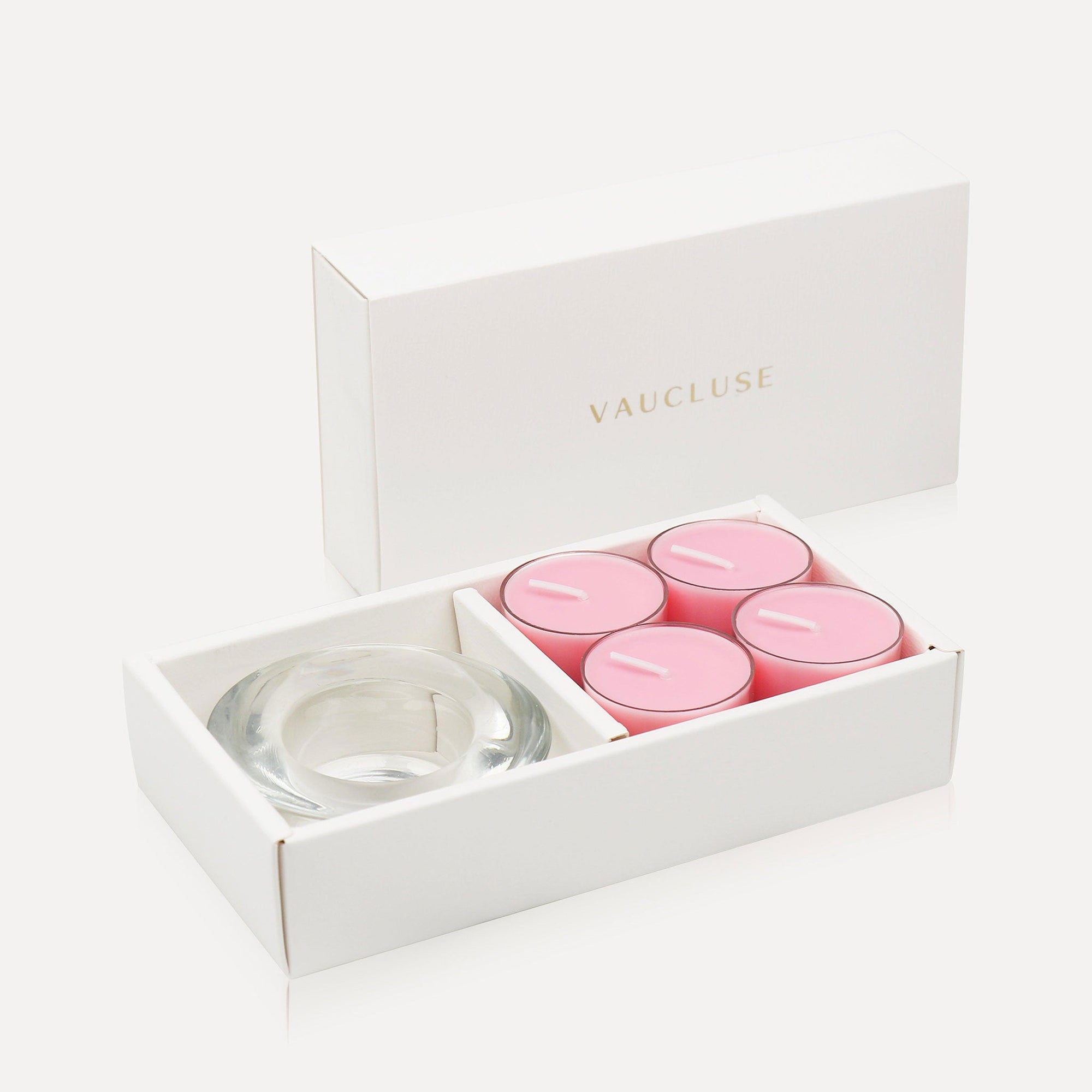 An open premium white gift box displaying four soft pink rose tealight candles alongside a thick round clear glass candle holder.