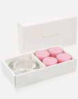 An open premium white gift box displaying four soft pink rose tealight candles alongside a thick round clear glass candle holder.