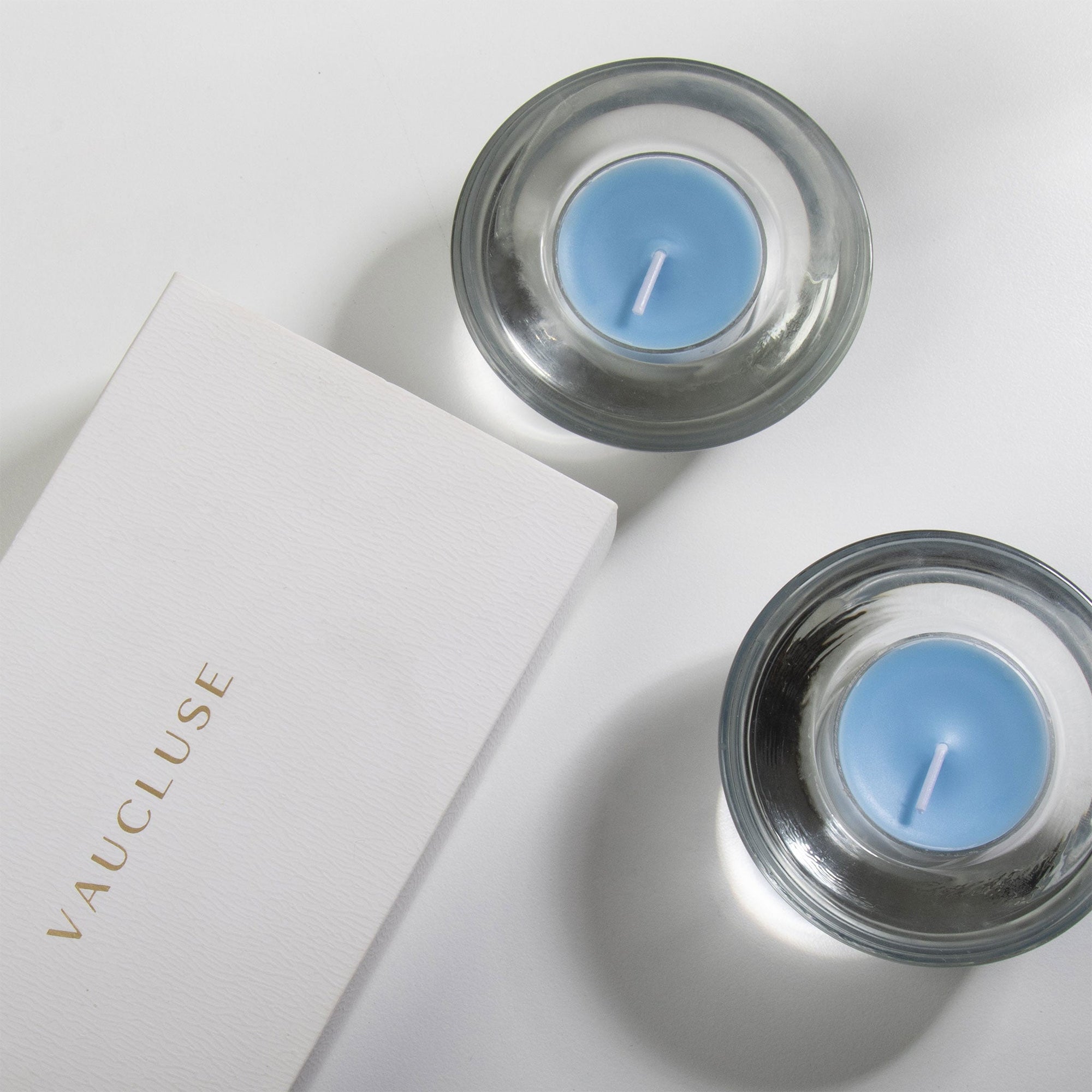 An overhead top-down view of two light blue sea breeze tealight candles resting in modern clear glass holders next to a white embossed Vaucluse gift box.