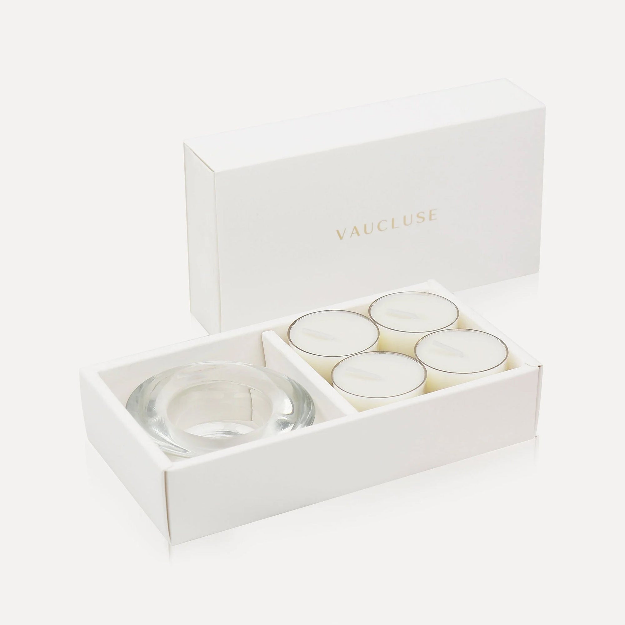 An open luxury white gift box displaying four premium vanilla tealight candles alongside a matching thick clear glass candle holder.