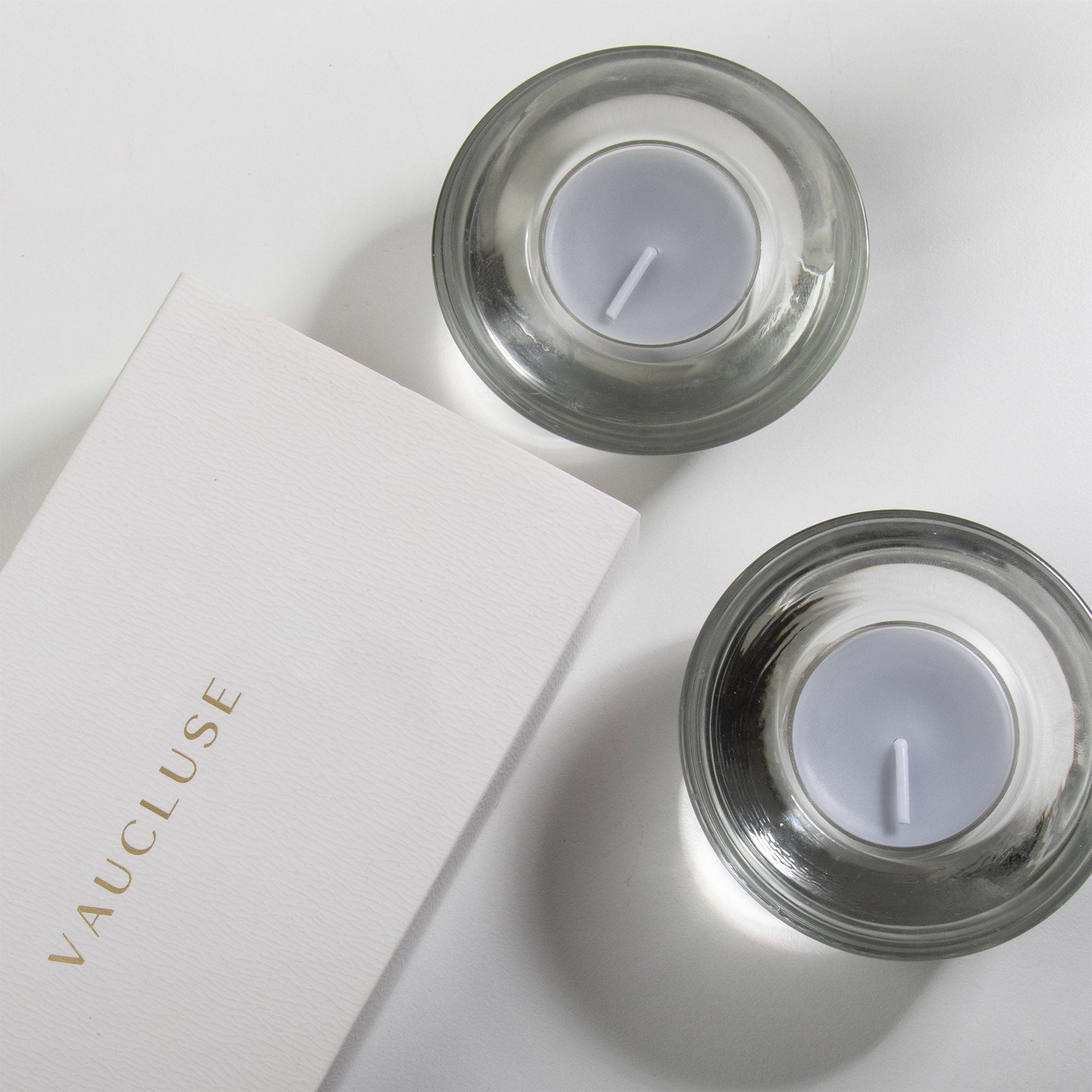 An overhead top-down view of two grey blue velvet tealight candles resting in modern clear glass holders next to a white embossed Vaucluse gift box.