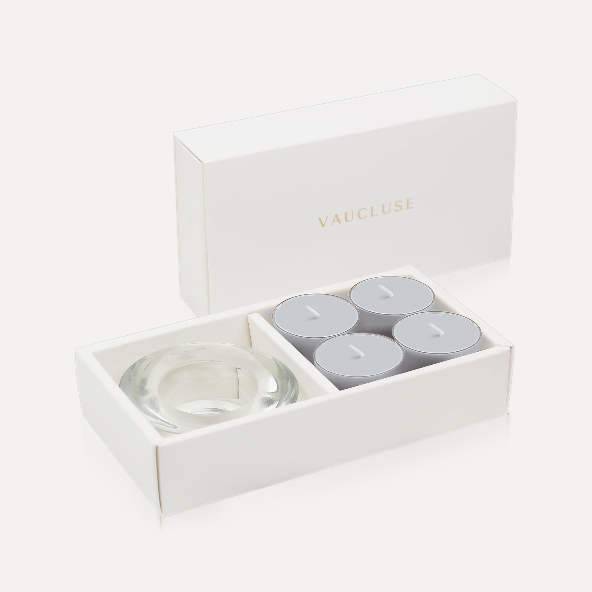 An open luxury white gift box displaying a set of four soft grey velvet tealight candles next to a round clear glass candle holder.