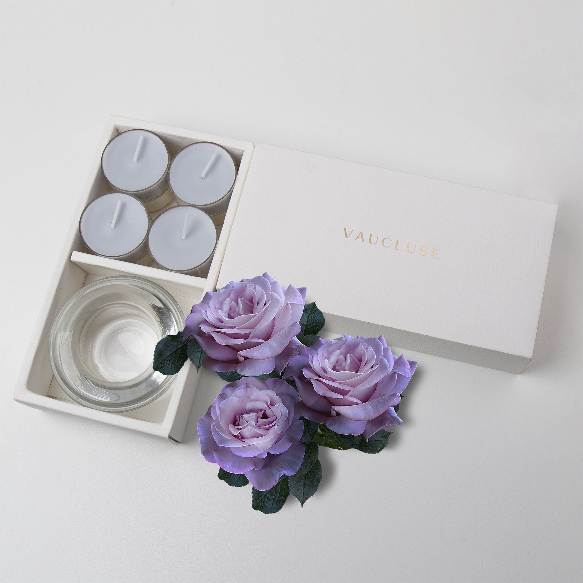 Minimalist top-down flat lay of the open velvet tealight candle set beautifully accessorized with three dusty purple rose blossoms on a white surface.