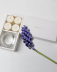 Minimalist overhead flat lay of the open white musk tealight candle set styled beautifully with a single purple hyacinth flower branch on a white background.